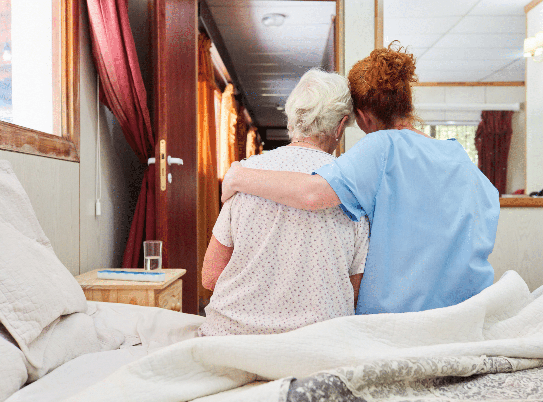 A nurse sits beside an elderly woman on a bed, providing care and companionship in a warm, supportive environment.