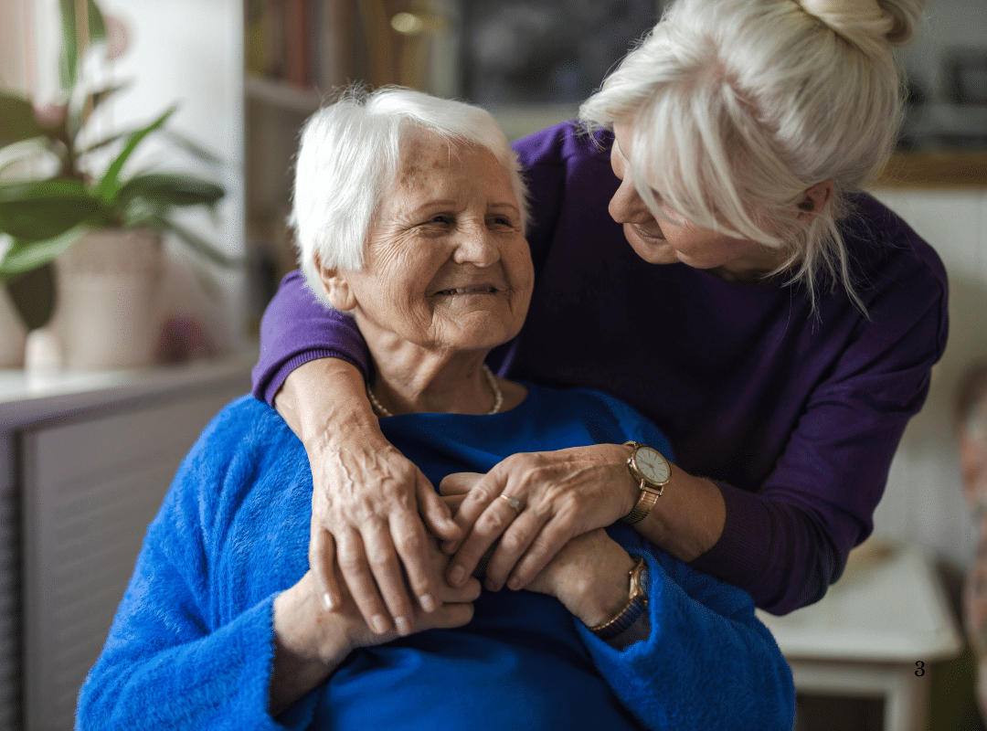Senior woman in blue sweater smiling warmly, embraced by caregiver in purple top, showcasing connection and care in assisted living environment.